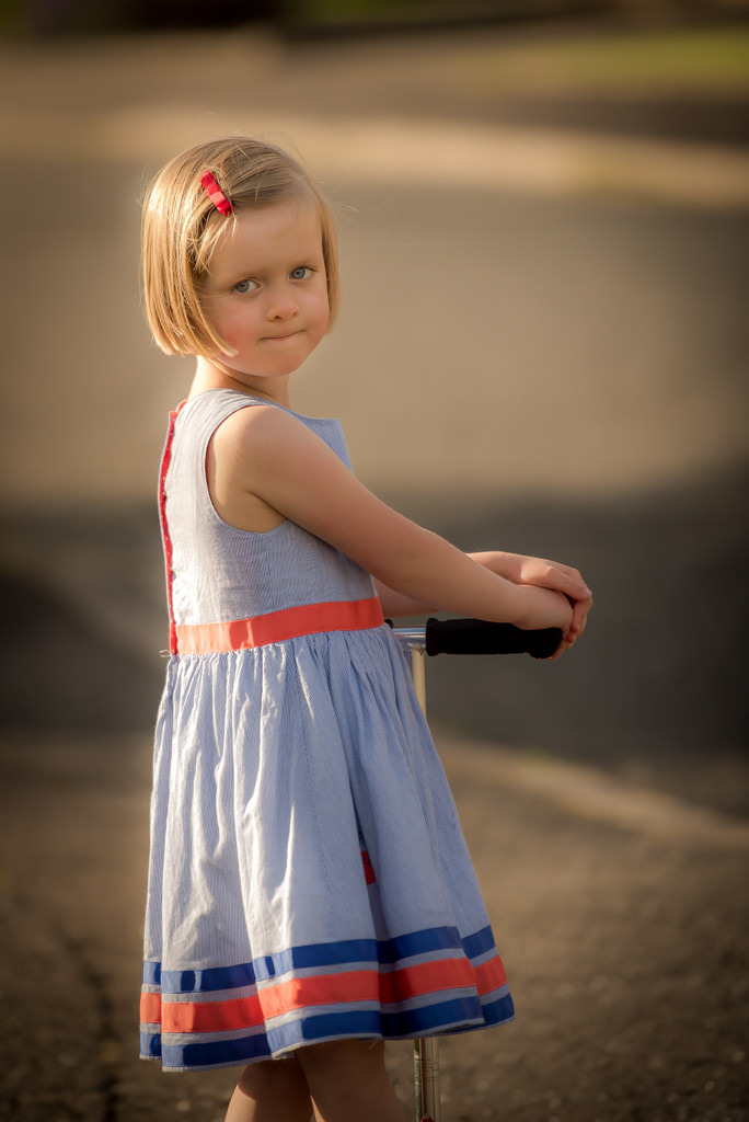 Family photographer Edinburgh - 4 year old girl with blonde hair and blue eyes wearing a red and blue dress standing on a scooter in the evening sunshine