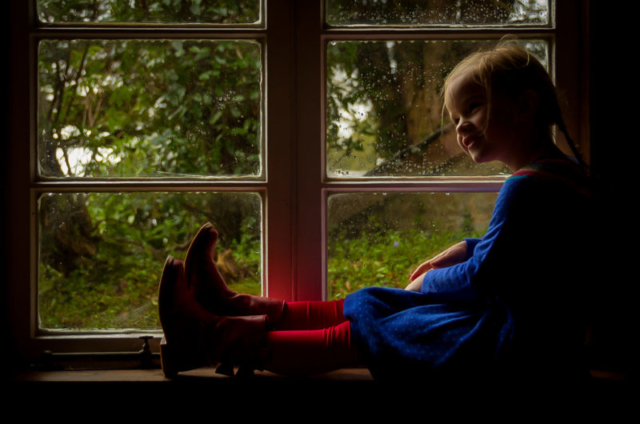 Family photographer Edinburgh - little girl in blue dress and red tights sitting in a window with rain outside