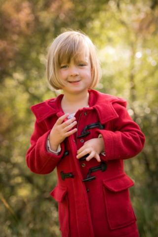 Family photographer Edinburgh - 4 year old girl, blonde hair and blue eyes, wearing bright red coat in the woods in the evening sunshine