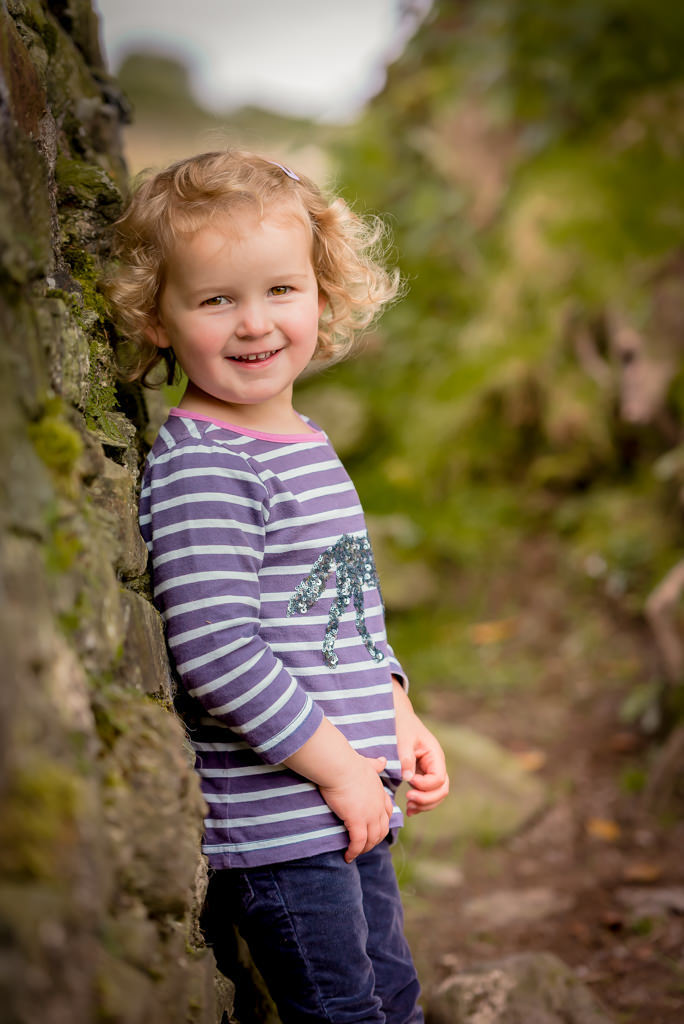 Family photographer Edinburgh - toddler girl with blonde curls leaning against a mossy dry stone wall