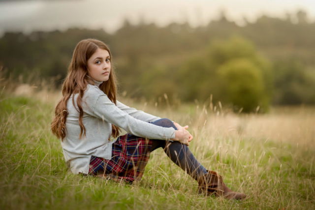 Family photographer Edinburgh - 12 year old girl in tartan skirt sitting in a field of grass