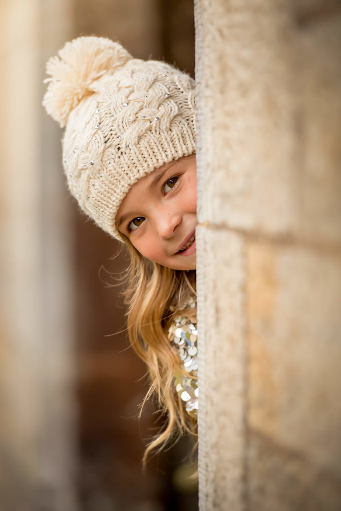 Family photographer Edinburgh - headshot of little girl with cream bobble hat on peeking round corner