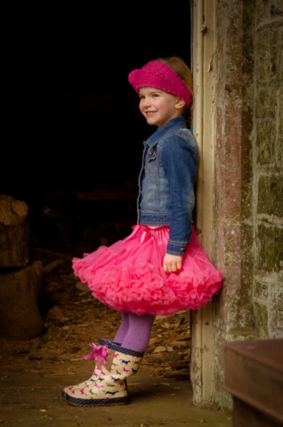 Family photographer Edinburgh - 4 year old girl with bright pink tutu, denim jacket, wellies, and pink hair band leaning in doorway of a barn