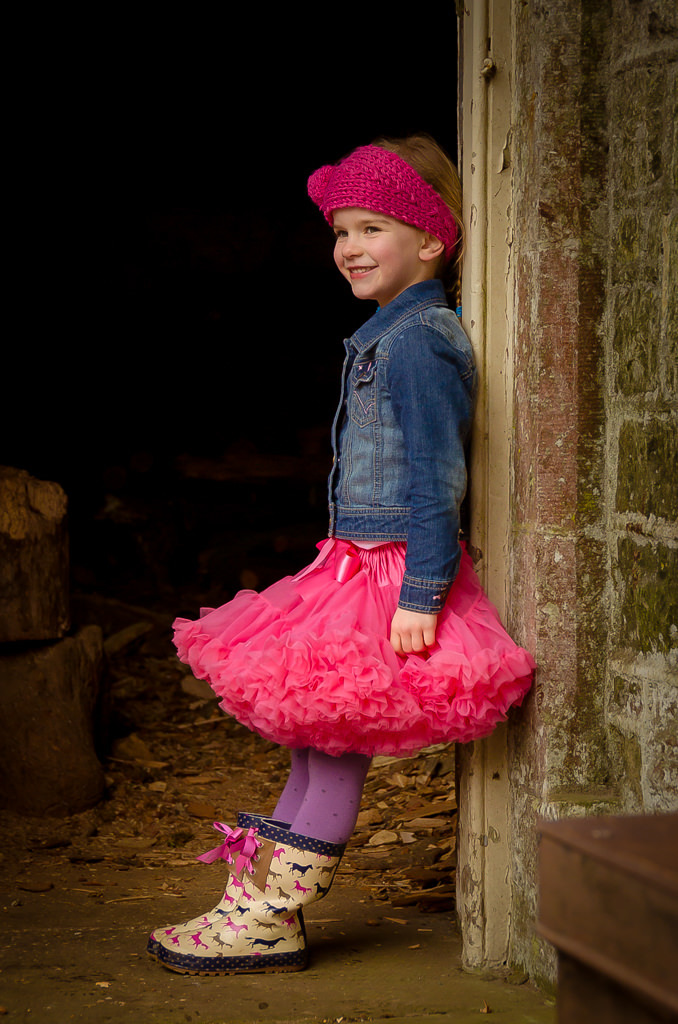 Family photographer Edinburgh - 4 year old girl with bright pink tutu, denim jacket, wellies, and pink hair band leaning in doorway of a barn