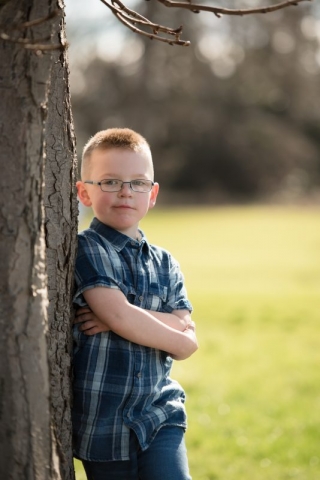 Family photographer Edinburgh - little boy in blue check shirt with glasses, leaning against a tree with arms folded