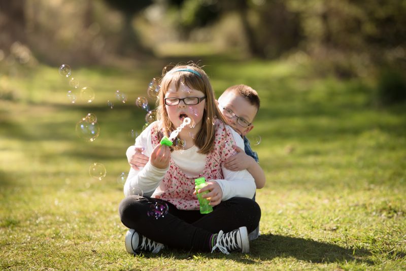 sunset photography sessions - Family photographer Edinburgh - little girl with glasses sitting on grass cross-legged blowing bubbles, brother peeking out behind her