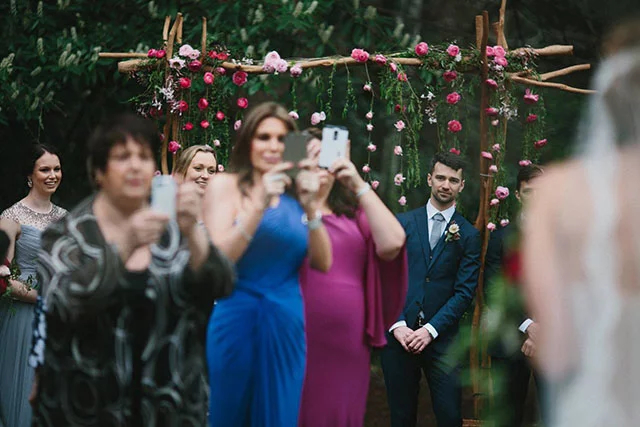 Bridegroom leaning past guests at his wedding with smartphones so he can see his bride walking up the aisle