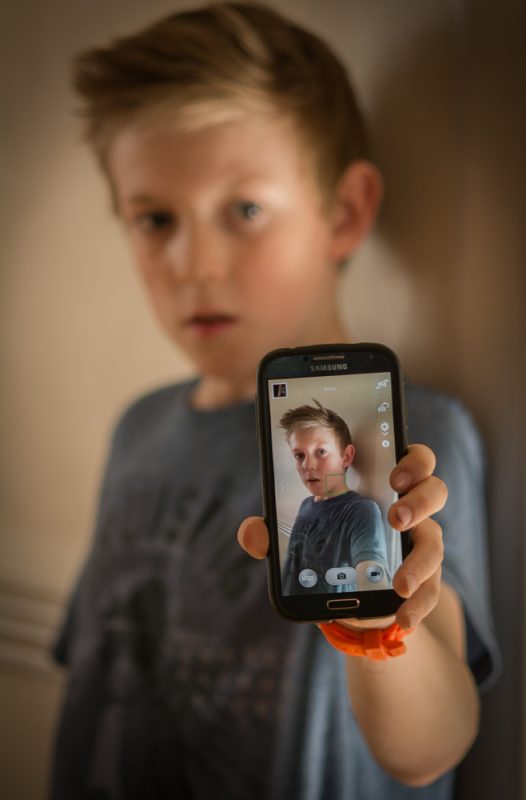 Smartphone addiction - young boy holding up mobile phone with selfie on it