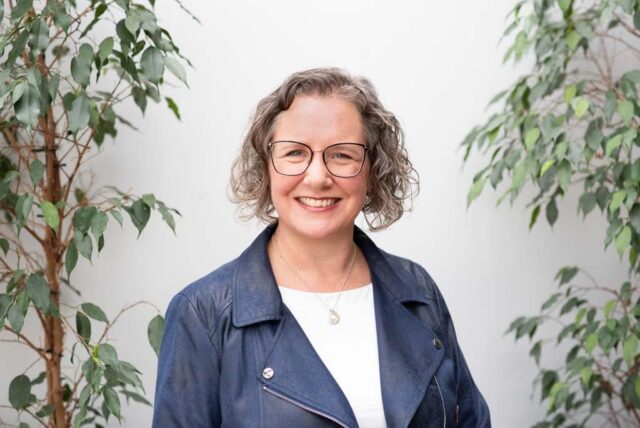 Smiling author and business consultant in a navy jacket, framed by green plants during an Edinburgh headshots and brand photography session.