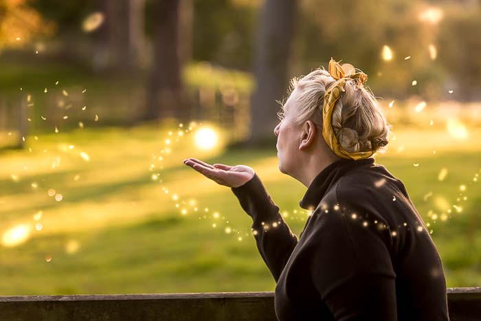 Brand photography in Edinburgh, Scotland, for small business owner showing woman with blonde hair and black jumper  in a woodland setting blowing pixie dust out of her hand