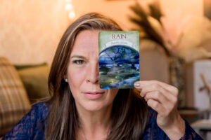 Headshot in Edinburgh, Scotland, of woman with long brown hair holding up an Oracle card in front of her eye